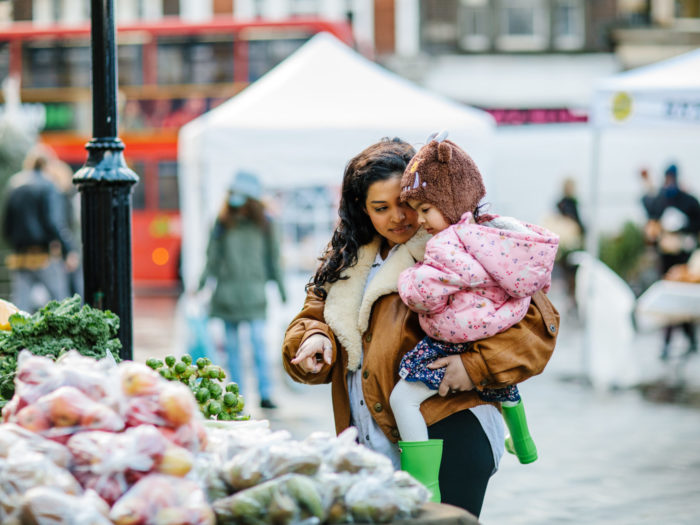 Woman holds child in her arms, and points at produce on a market stall.