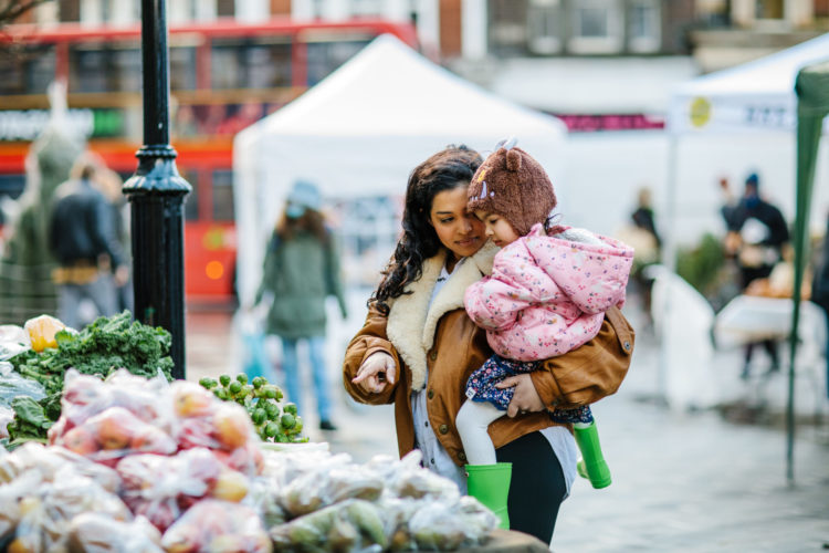 Woman holds child in her arms, and points at produce on a market stall.