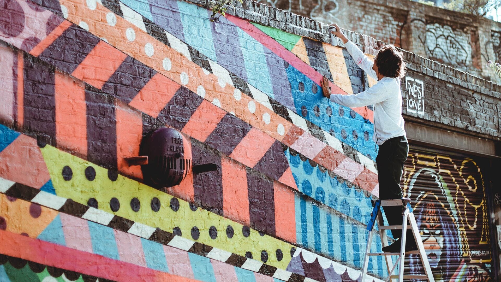 Graffiti wall, with person standing on ladder and reaching over the wall. Photo by Abi Ismail