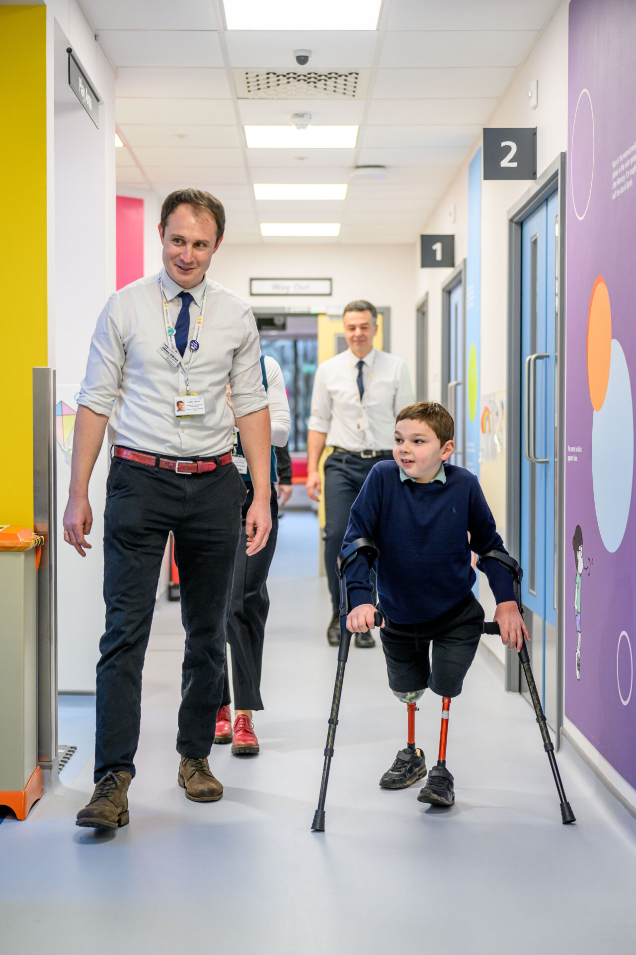 A clinician walks along a corridor with a child using crutches and leg prosthetics, at the Evelina Day Centre